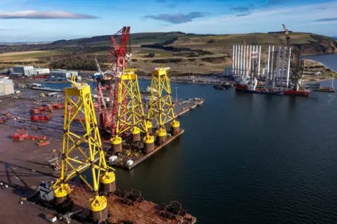 Port of Nigg The Port of Nigg is a large industrial area on the Moray Firth. On its quaysides are yellow structures for offshore wind farms called jackets.
/p
pAcross an area of water from the jackets are white offshore wind turbine blades lined up in a row.