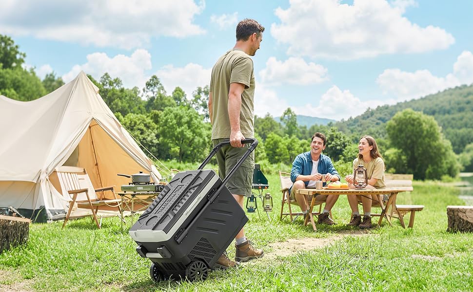 Person rolling wheeled case across grassy campground with large bell tent and picnic table in background. Scenic mountain landscape with trees visible.
