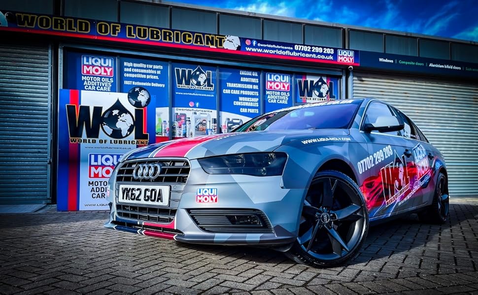 Silver Audi vehicle parked in front of a storefront with blue lighting at night. Custom wheels and promotional vehicle wrap visible.