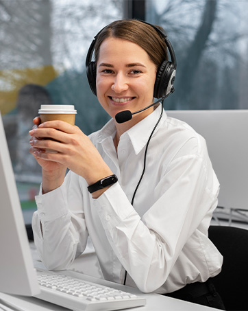 Person wearing headset at desk with laptop, holding coffee cup. White shirt, office setting visible.
